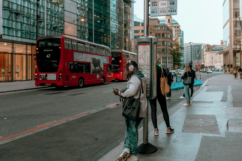 A busy street with people waiting at a bus stop and red double-decker buses passing by.